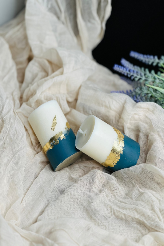 Two cylindrical candles with blue and gold accents on a textured fabric surface - UK made